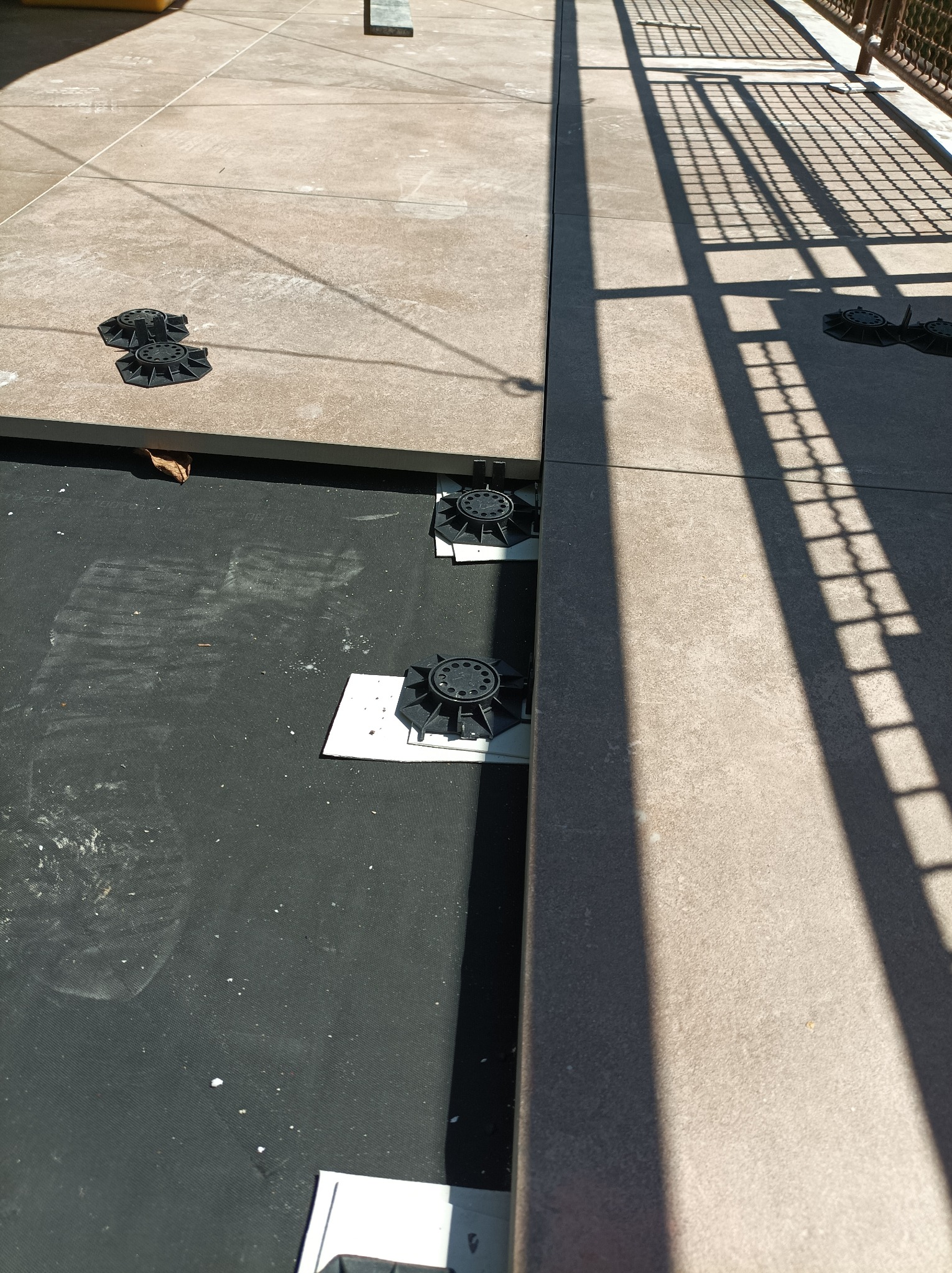 Montaż tarasu wentylowanego: widoczne czarne regulowane wsporniki na membranie hydroizolacyjnej, częściowo ułożone płyty tarasowe i cień metalowej balustrady.