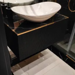 Nowoczesna łazienka z czarno-białymi płytkami marmurowymi, białą umywalką nablatową o organicznym kształcie i czarną szafką z ryflowanymi frontami oraz złotymi akcentami.