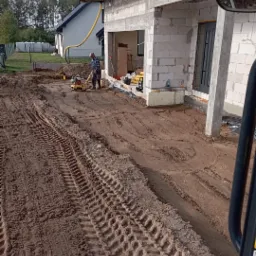 Prace ziemne przed domem w trakcie budowy: ubijanie gruntu zagęszczarką, widoczne ślady opon maszyny budowlanej, częściowo wykończona elewacja z betonu komórkowego.