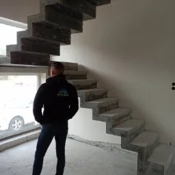 Surowe betonowe schody wewnętrzne w trakcie budowy, widok z dołu na konstrukcję, z pracownikiem w bluzie z logo firmy budowlanej stojącym w pobliżu okna.