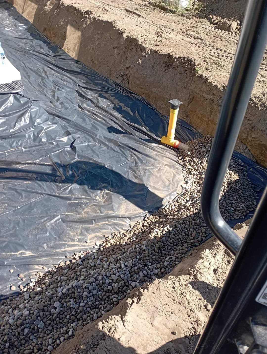 Wykop wyłożony czarną folią, wypełniony drobnym żwirem, z widoczną żółtą rurą drenażową i fragmentem koparki w kadrze.