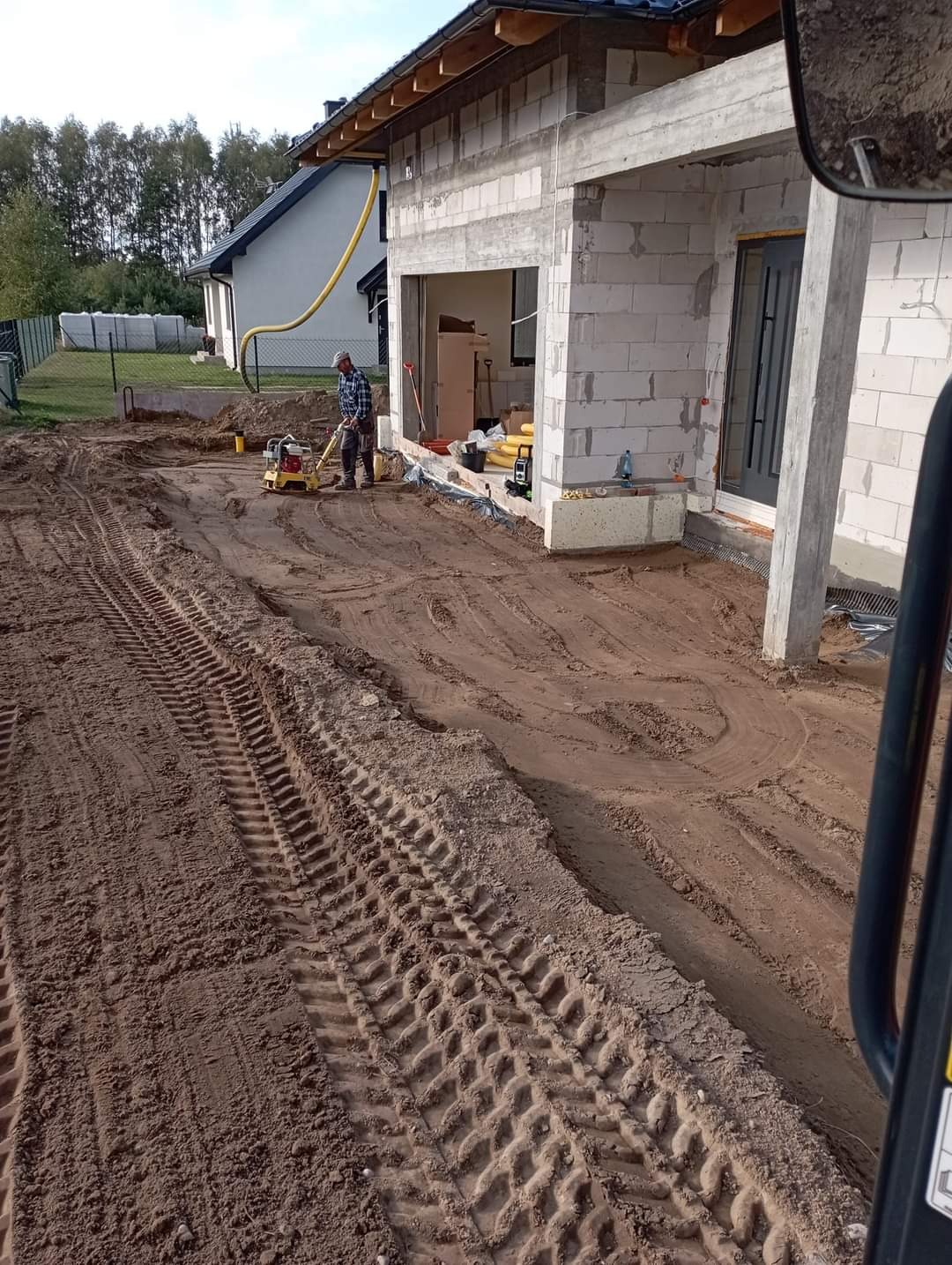 Prace ziemne przed domem w trakcie budowy: ubijanie gruntu zagęszczarką, widoczne ślady opon maszyny budowlanej, częściowo wykończona elewacja z betonu komórkowego.