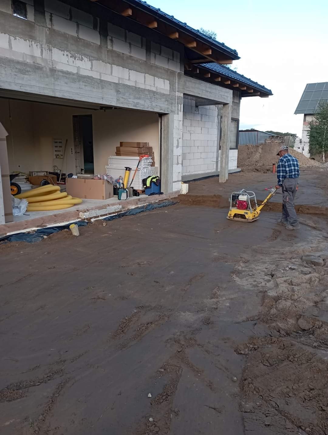 Pracownik w kraciastej koszuli zagęszcza ubijarką wibracyjną grunt przed budynkiem w stanie surowym otwartym z bloczków betonowych, widoczne przygotowane rury drenażowe.