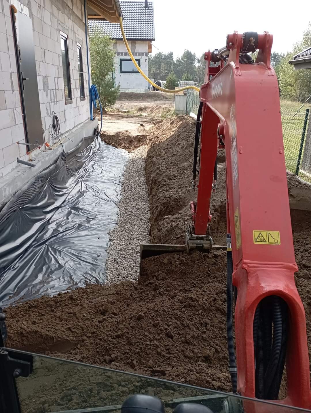 Czerwona minikoparka Kubota w Świeciu podczas prac ziemnych przy budowie domu, widoczny wykop zabezpieczony czarną folią i wypełniony żwirem, przygotowany pod fundament.