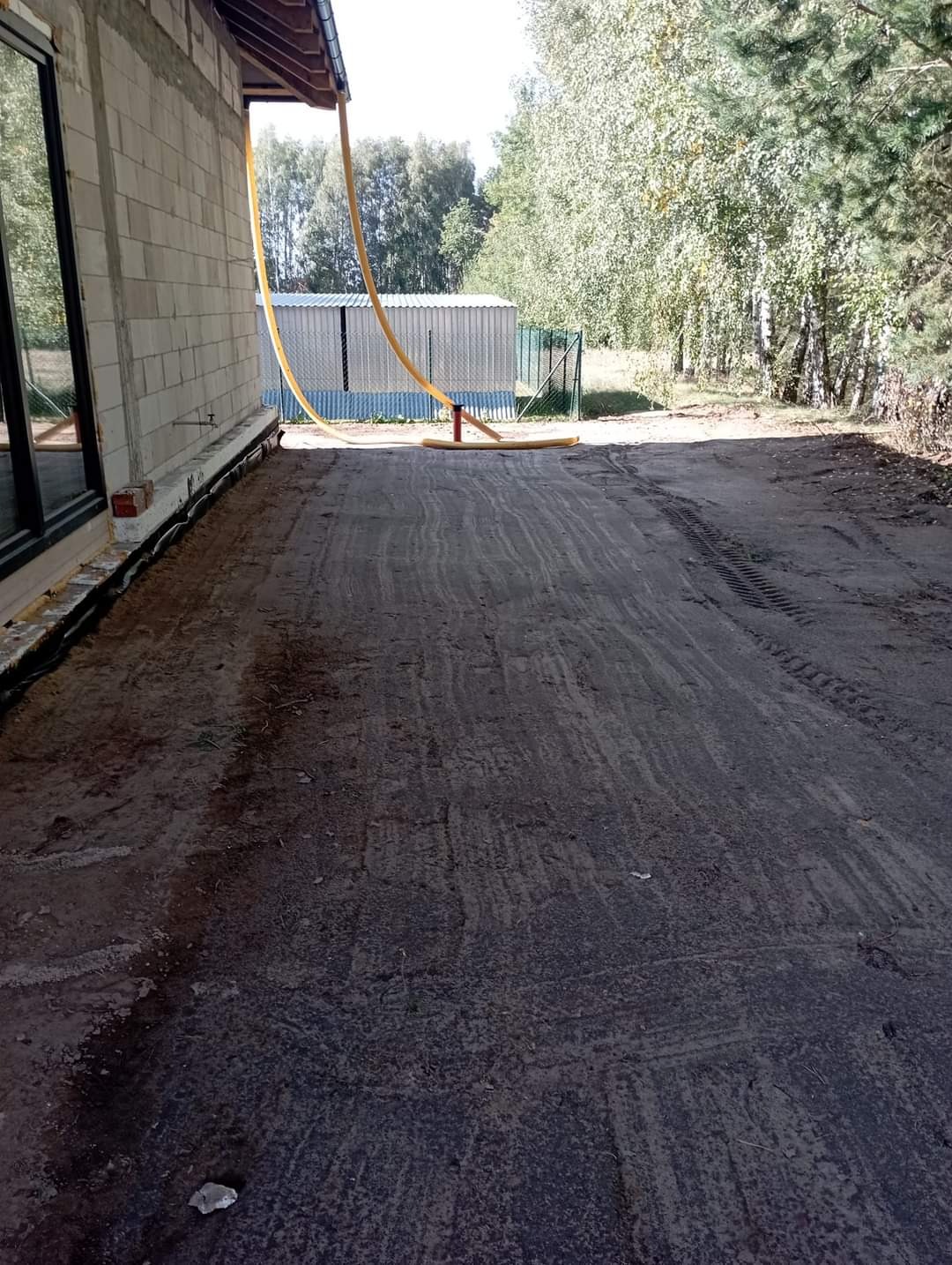 Teren po deszczu obok surowego domu z pustaków, z żółtym wężem odprowadzającym wodę z dachu, widoczne ślady opon na mokrej ziemi.