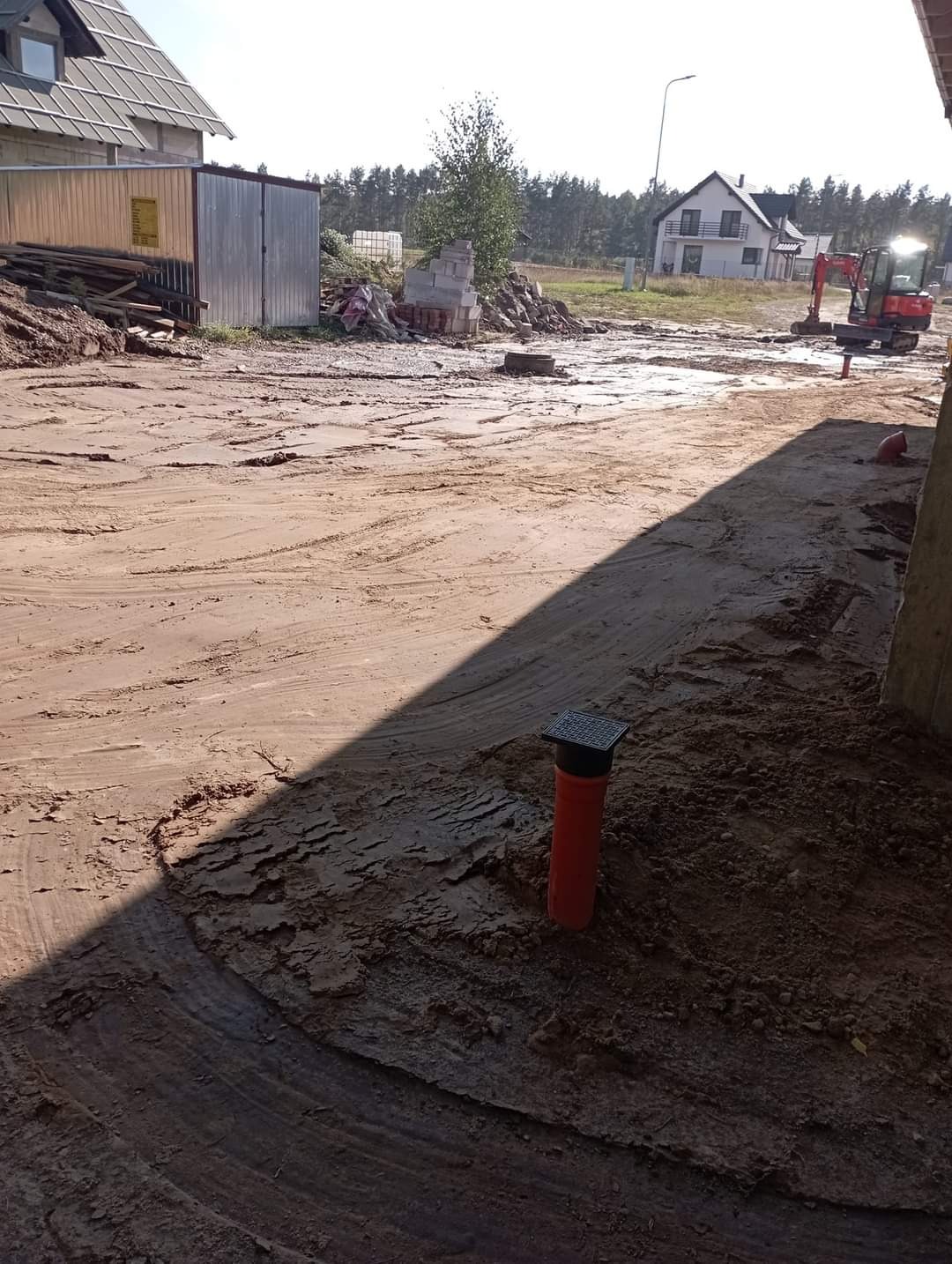 Pomarańczowa rura drenażowa wystająca z ziemi na placu budowy, widoczny wykopany teren, w tle dom w budowie i mini koparka.