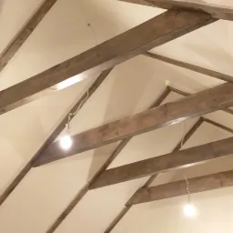 Pomalowane na kremowo poddasze z widocznymi drewnianymi belkami stropowymi i wiszącymi żarówkami na kablach.