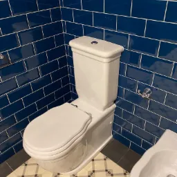 Biała toaleta z bidetem na wzorzystej podłodze, otoczona błyszczącymi, granatowymi płytkami ułożonymi w cegiełkę z jasną fugą i chromowanymi wieszakami na ścianie.