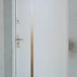 Białe drzwi wejściowe z pionowym, srebrnym paskiem, zamontowane w szarej, otynkowanej ścianie, widoczne klamka i wkładka zamka, z wycieraczką u dołu.