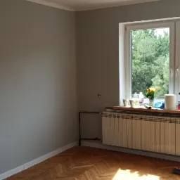 Pokój po remoncie: ściany pomalowane na szaro, drewniany parkiet, okno z widokiem na zieleń, na parapecie kwiaty i akcesoria malarskie, widoczna rura grzejnika i stojąca miotła.