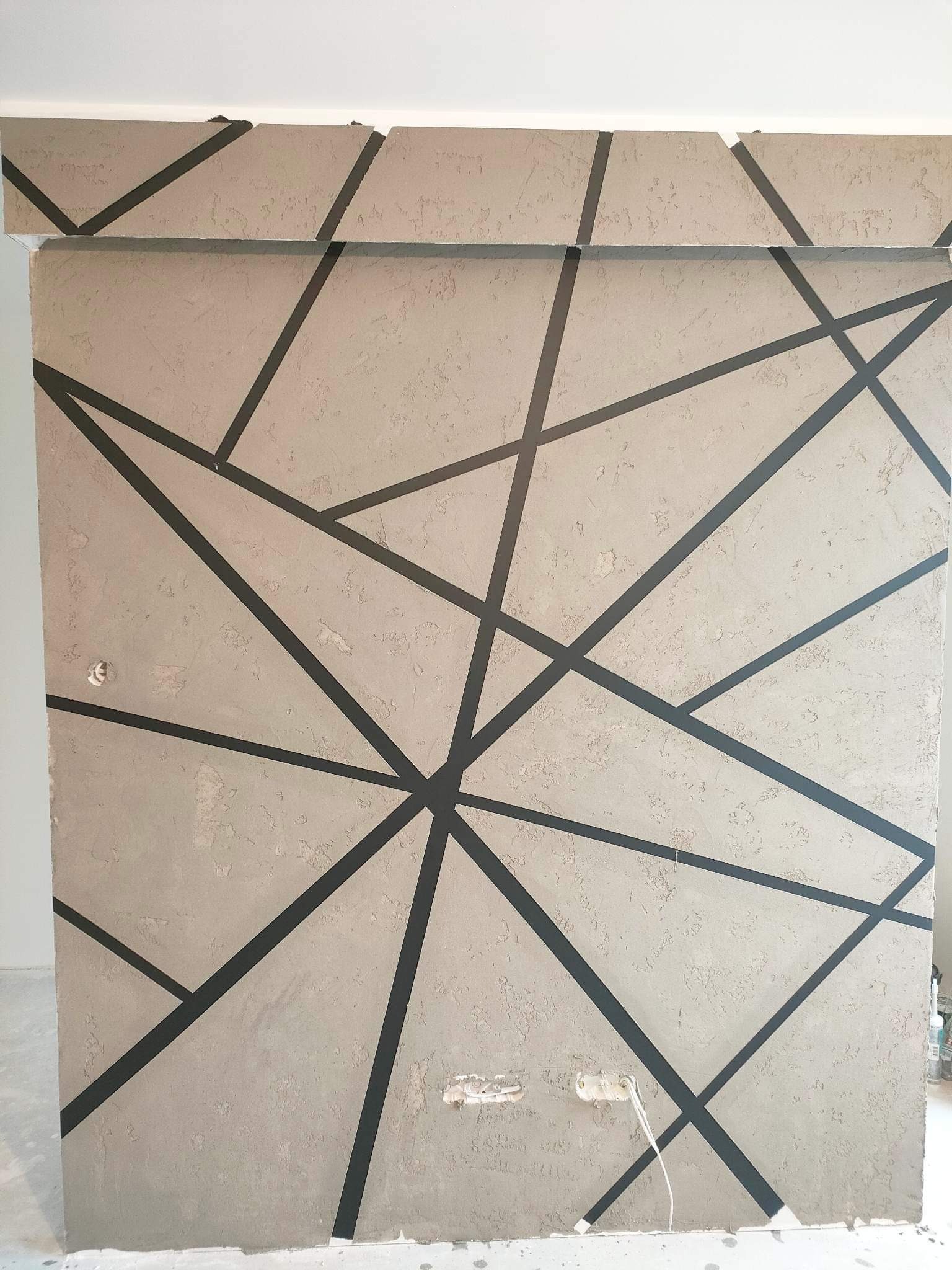 Ściana z dekoracyjną, geometryczną strukturą w Radomsku, wykonana z szarego tynku i czarnych pasów, widoczne niedoskonałości wykończenia i gniazdko elektryczne.