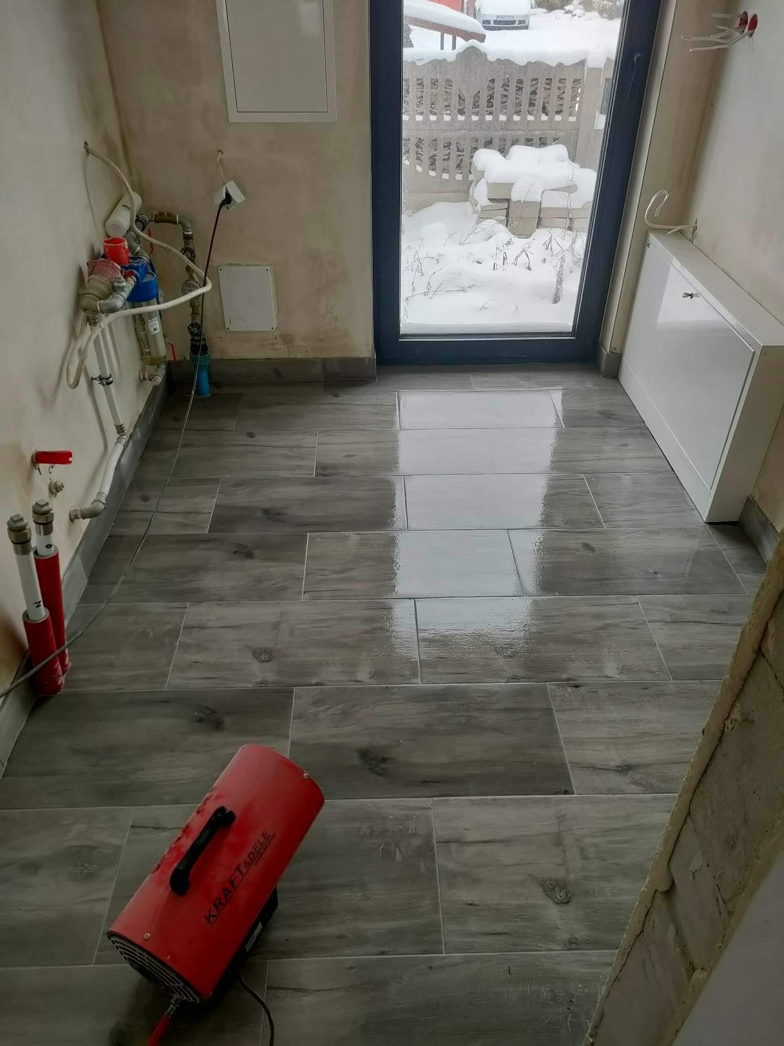 Ułożona podłoga z szarych paneli imitujących drewno w pomieszczeniu z widoczną instalacją hydrauliczną i nagrzewnicą powietrza, za oknem śnieg.