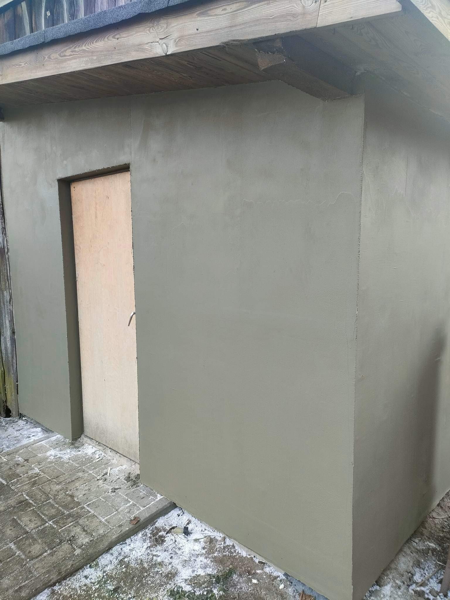 Otynkowana, szara, mała budowla z drewnianym dachem i surowymi, drewnianymi drzwiami, widoczna z zewnątrz, z fragmentem kostki brukowej na dole.