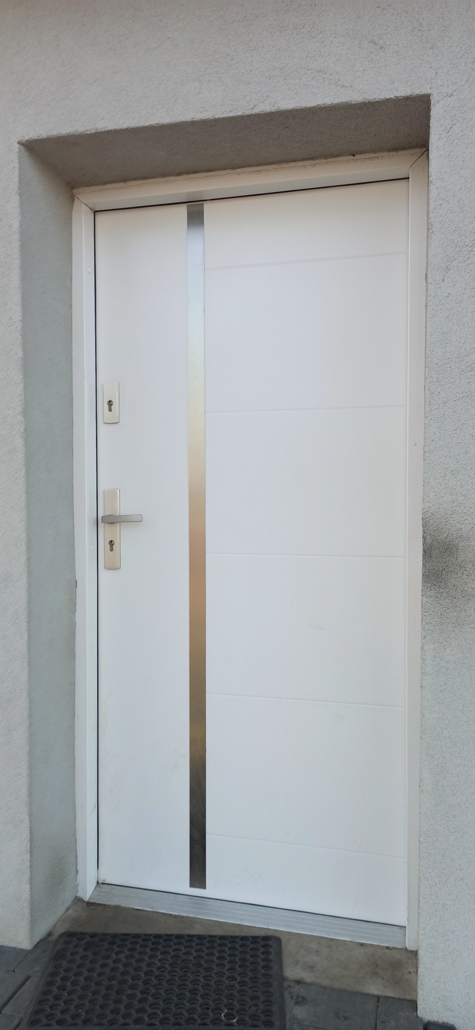 Białe drzwi wejściowe z pionowym, srebrnym paskiem, zamontowane w szarej, otynkowanej ścianie, widoczne klamka i wkładka zamka, z wycieraczką u dołu.