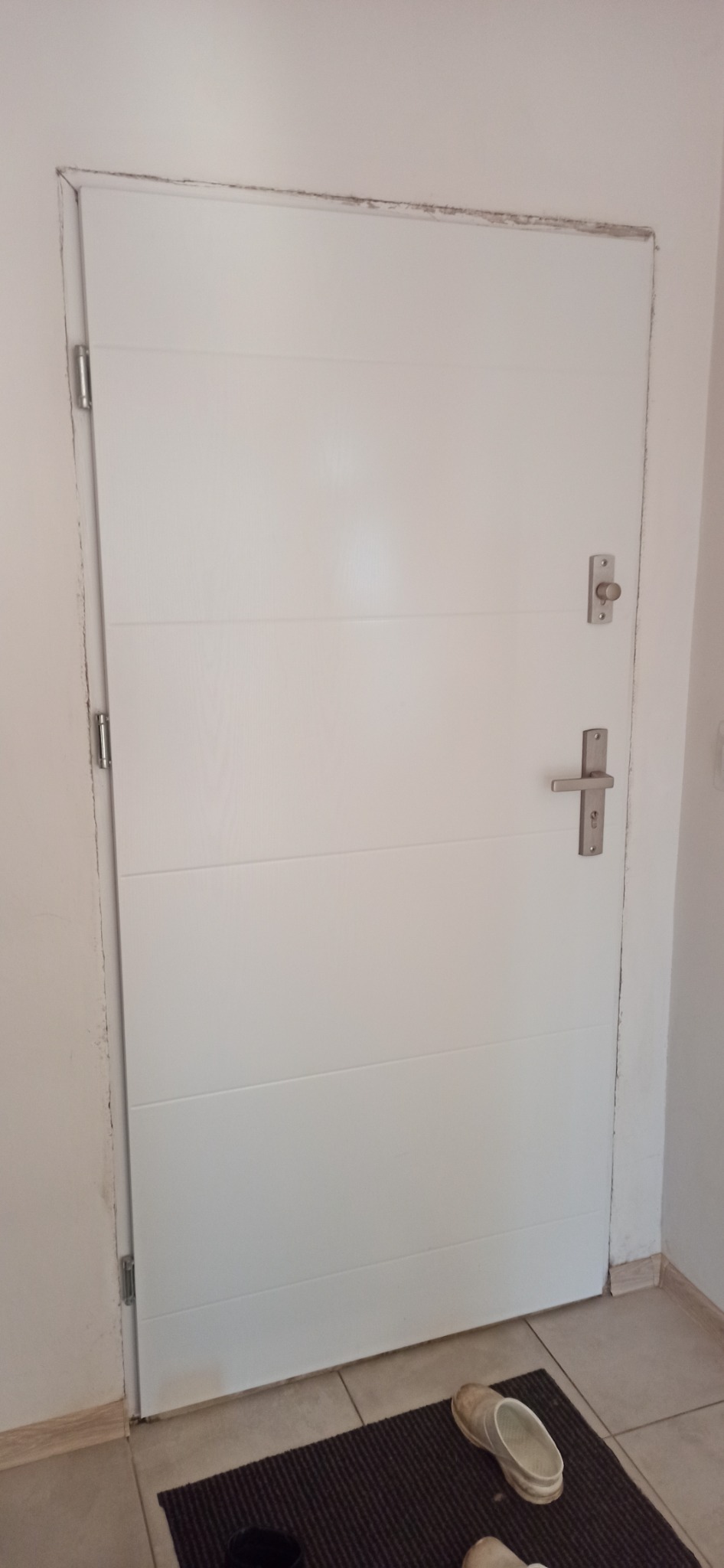 Białe, poziomo żłobione drzwi wewnętrzne z metalową klamką i zamkiem, widoczne ślady po malowaniu na ścianie i ościeżnicy, brudna wycieraczka i obuwie w dolnej części kadru.