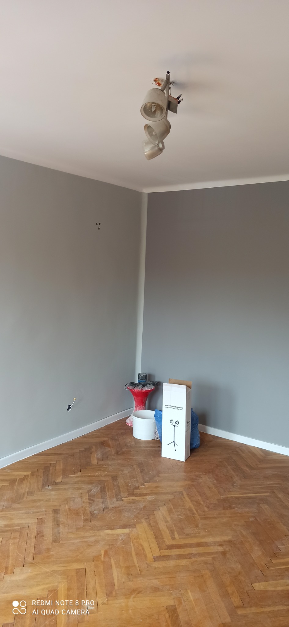 Pokój po malowaniu ścian na szaro, widoczna drewniana podłoga w jodełkę, puszka z farbą i karton z lampą halogenową w rogu.