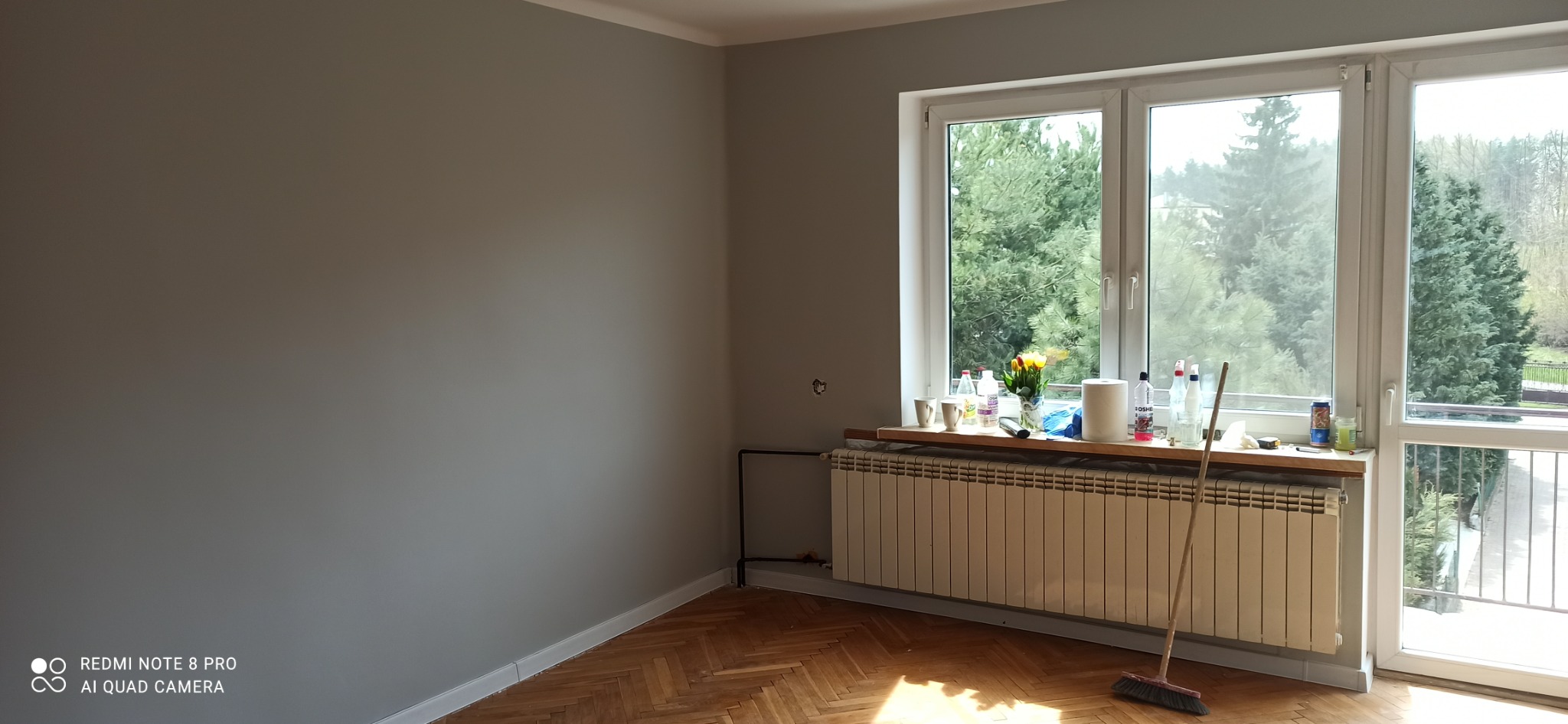 Pokój po remoncie: ściany pomalowane na szaro, drewniany parkiet, okno z widokiem na zieleń, na parapecie kwiaty i akcesoria malarskie, widoczna rura grzejnika i stojąca miotła.