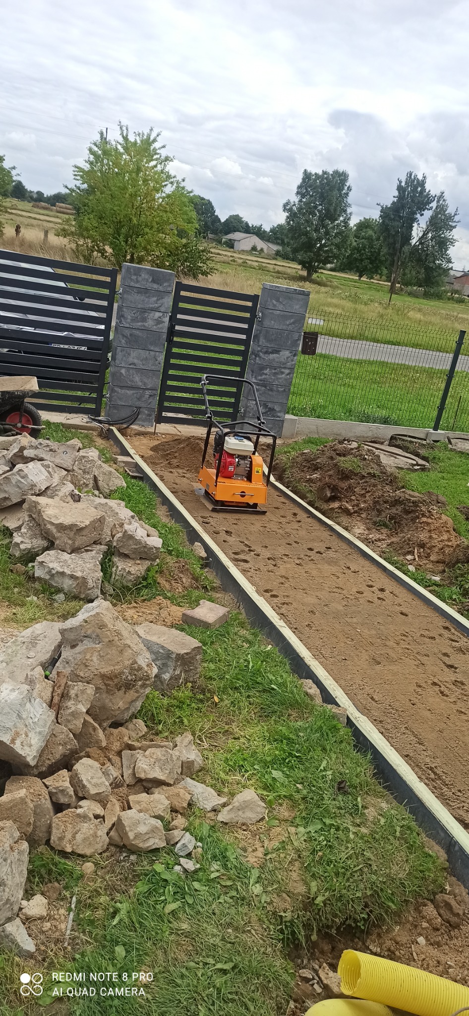 Pomarańczowa zagęszczarka do gruntu stoi na piasku pomiędzy obrzeżami, przygotowując podłoże pod chodnik z kostki, widoczne kamienie, brama i trawa.
