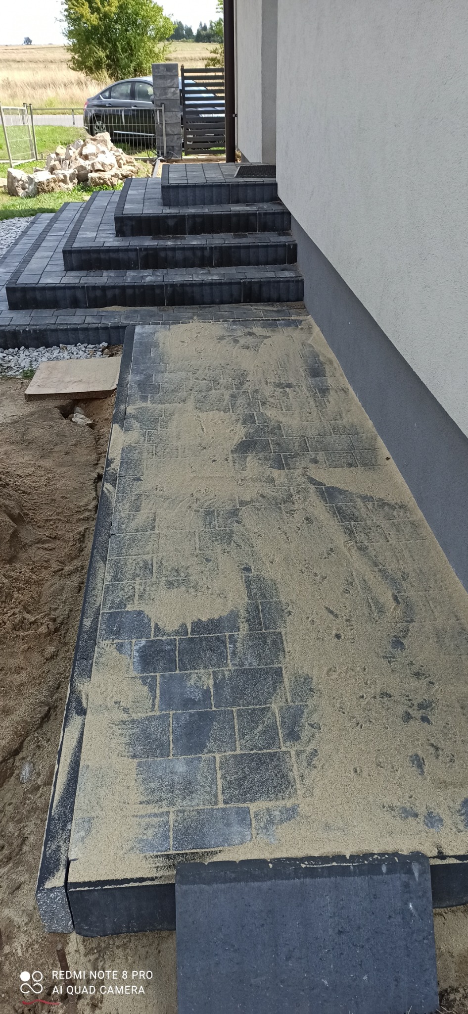 Ułożona, szara kostka brukowa na podjeździe i schodach wejściowych do domu, posypana piaskiem w celu wypełnienia szczelin, widoczny fragment elewacji i samochód w tle.