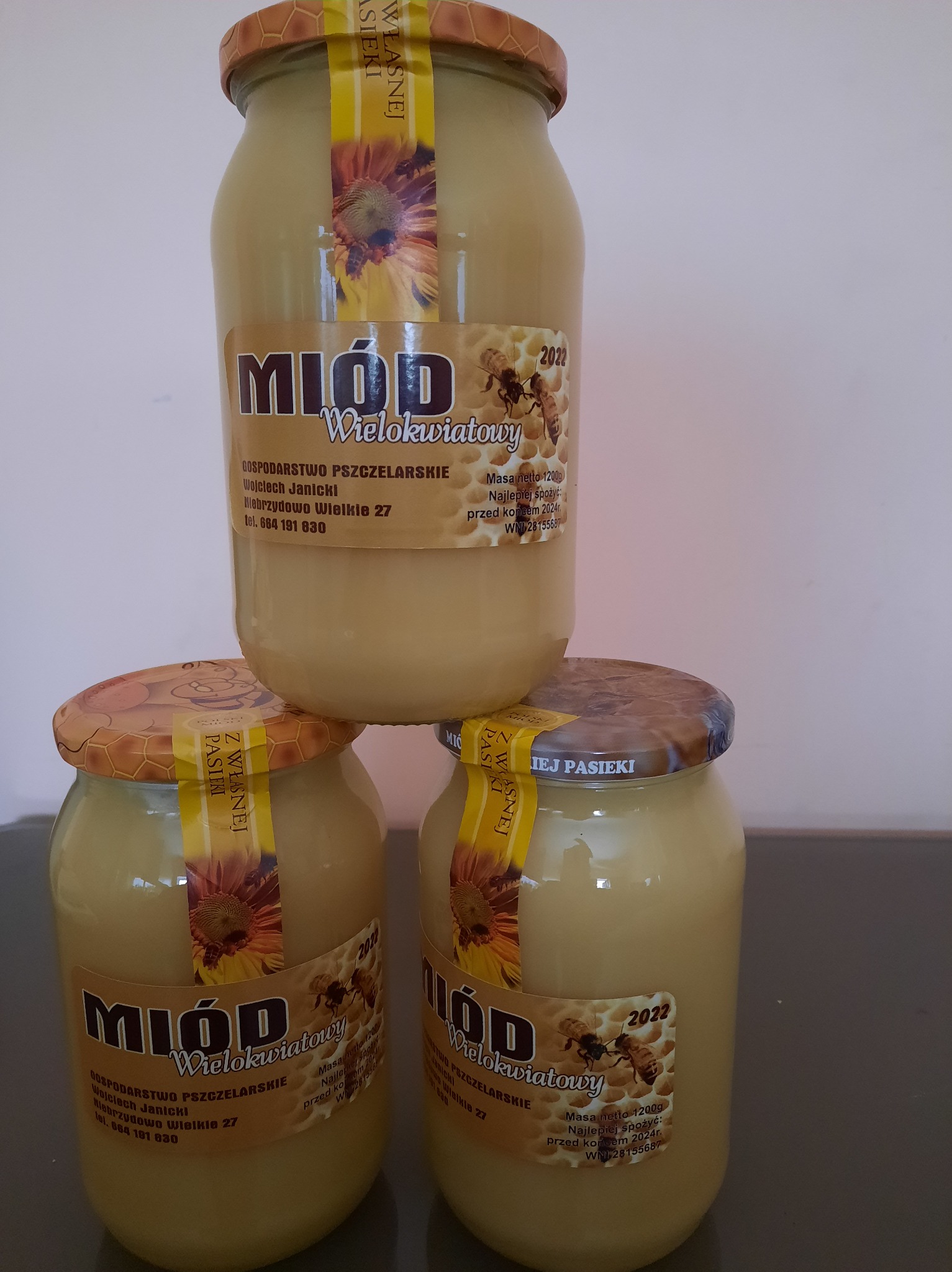 Trzy słoiki wielokwiatowego miodu z pasieki Wojciecha Janickiego, ułożone jeden na drugim, z widocznymi etykietami informującymi o dacie przydatności do spożycia i masie netto.