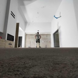 Remonty i Wykończenia Bartłomiej Kirejczyk - Wysoki salon w trakcie wykończenia: gładzie na ścianach, zabudowa kominka, instalacje elektryczne. Widok z niskiej perspektywy na majstra z rękami na biodrach.