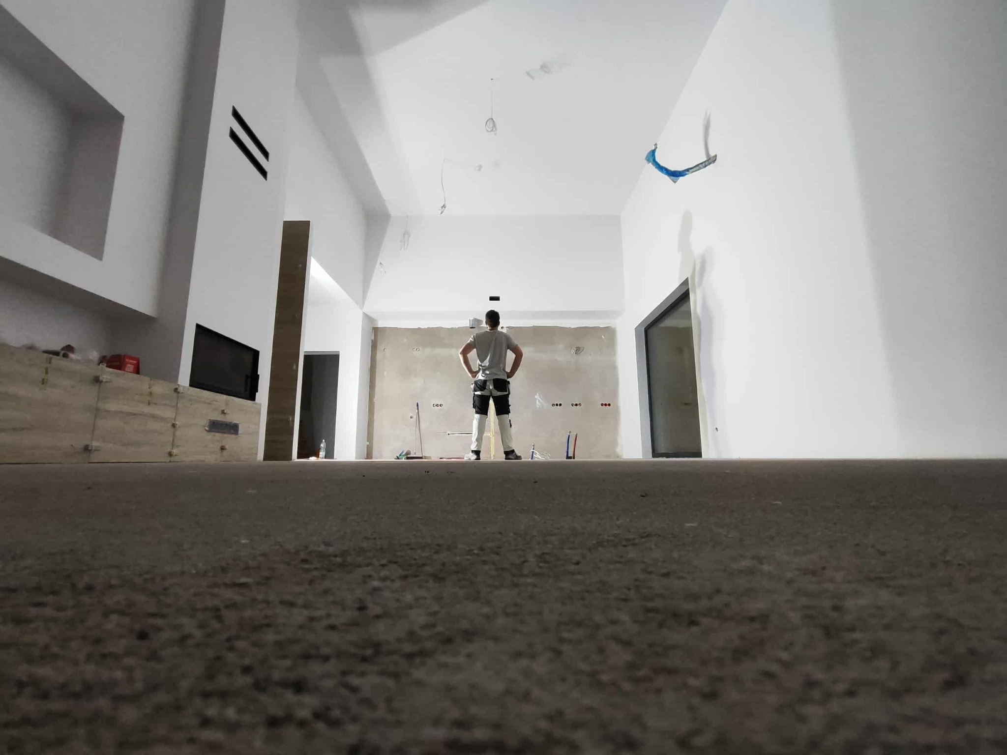 Wysoki salon w trakcie wykończenia: gładzie na ścianach, zabudowa kominka, instalacje elektryczne. Widok z niskiej perspektywy na majstra z rękami na biodrach.