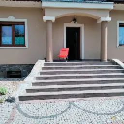 Wejście do domu w Bolesławcu z szerokimi schodami wyłożonymi płytkami imitującymi drewno, jasna elewacja z kolumnami i czerwonym krzesłem przed drzwiami.