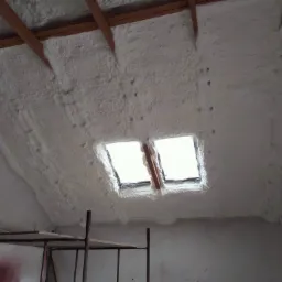 Poddasze po natrysku pianki poliuretanowej, widoczne okna dachowe i drewniana konstrukcja więźby dachowej, w tle rusztowanie.