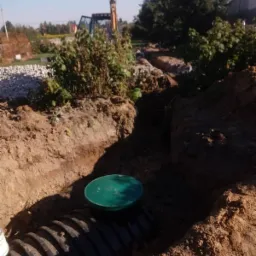 Czarna rura kanalizacyjna z zieloną pokrywą studzienki w wykopie ziemnym, w tle widoczna koparka i krzewy.