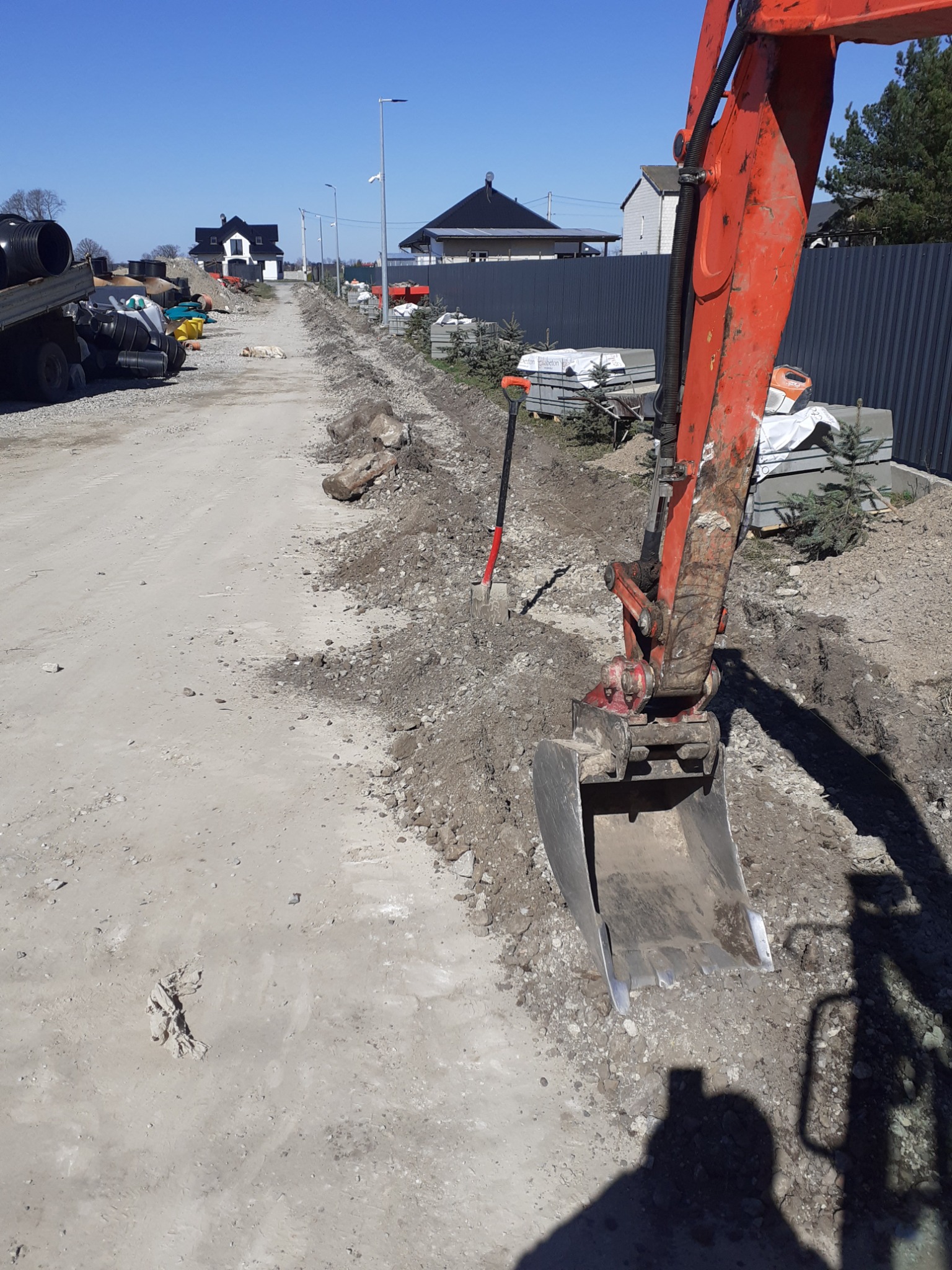 Pomarańczowa koparka przygotowuje teren pod przyszły chodnik, widoczny wykop, krawężniki i sadzonki drzewek, w tle domy jednorodzinne i szara metalowa siatka ogrodzeniowa.