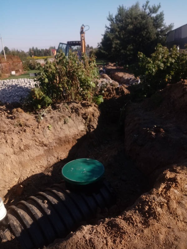 Czarna rura kanalizacyjna z zieloną pokrywą studzienki w wykopie ziemnym, w tle widoczna koparka i krzewy.