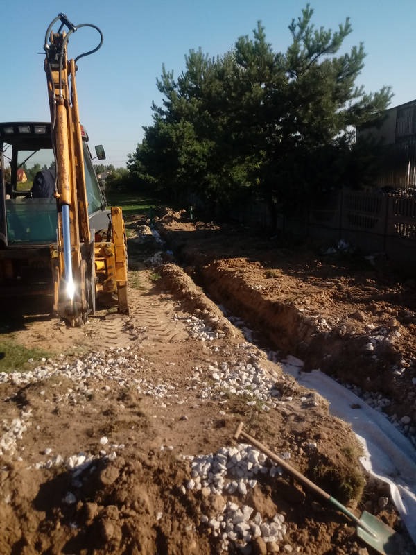 Żółta koparka stoi obok wykopanego rowu z ułożoną białą geowłókniną, zasypywanego drobnym żwirem, na tle drzew i niebieskiego nieba.