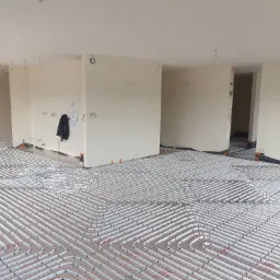 Rozległa instalacja ogrzewania podłogowego z białych rurek na czerwonej siatce, widok z góry na surowy stan wykończenia wnętrza, z widocznymi otworami na oświetlenie w suficie i elementami...