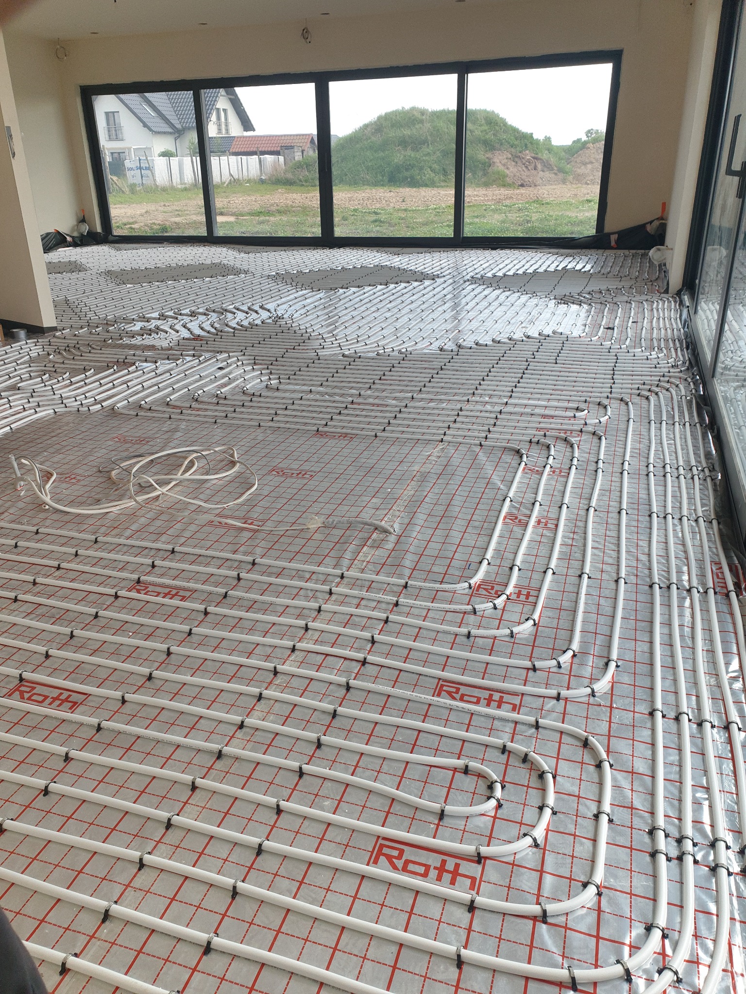 Rozłożone rury ogrzewania podłogowego marki Roth na folii z czerwoną kratką w jasnym pomieszczeniu z dużymi oknami.