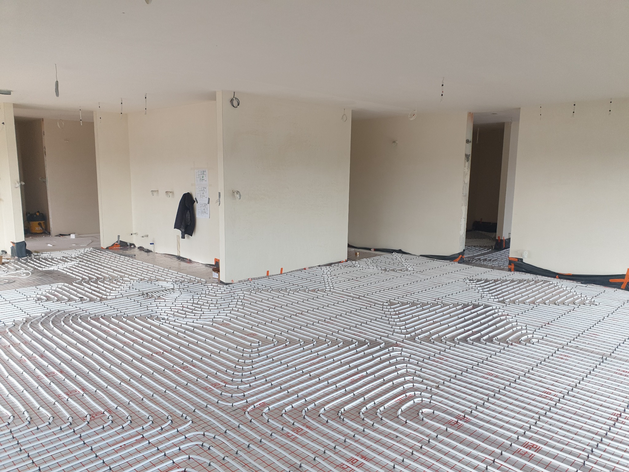 Rozległa instalacja ogrzewania podłogowego z białych rurek na czerwonej siatce, widok z góry na surowy stan wykończenia wnętrza, z widocznymi otworami na oświetlenie w suficie i elementami...