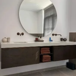 Nowoczesna łazienka z podwójną umywalką nablatową, okrągłym lustrem i ciemnymi, drewnianymi szafkami wiszącymi, uzupełniona o brązowe ręczniki i minimalistyczną armaturę w kolorze miedzi.