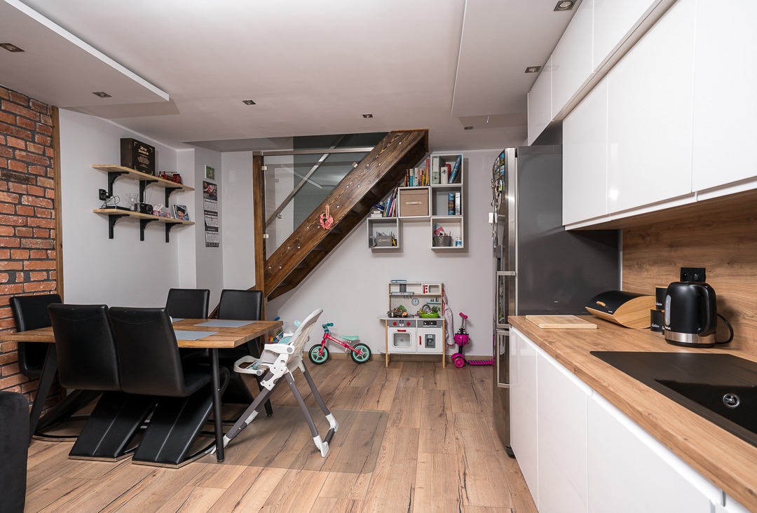 Jasne wnętrze domu z drewnianą podłogą, ścianą z cegły, schodami z przeszkloną balustradą, stołem z czarnymi krzesłami, białą kuchnią i dziecięcymi zabawkami.