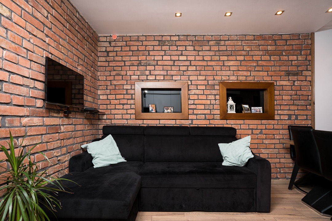 Ciemna, narożna sofa z jasnymi poduszkami na tle ściany z czerwonej cegły z wnękami z ramkami, w nowoczesnym salonie.
