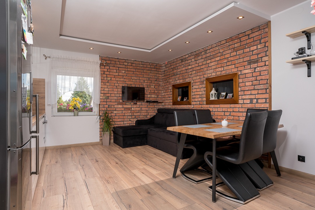 Salon z ceglaną ścianą, czarnym narożnikiem, dębowym stołem i krzesłami z czarnej ekoskóry, drewniana podłoga, okno z kwiatami w doniczce.