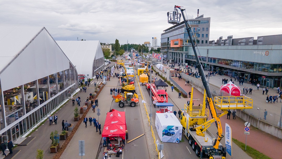 Widok z drona na targi firmowe z licznymi stoiskami, maszynami budowlanymi i pawilonami wystawienniczymi, z budynkiem AmberExpo w tle i grupą ludzi spacerujących po terenie.