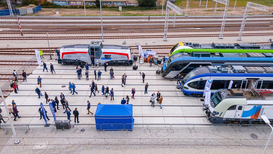 Z lotu ptaka: Prezentacja kilku nowoczesnych pociągów na tle torów kolejowych, otoczonych przez grupę ludzi i namiot.