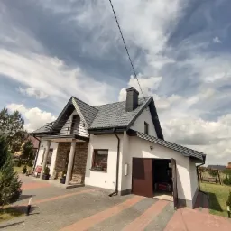 Dom jednorodzinny z szarym dachem z dachówki ceramicznej, widoczny komin i obróbki blacharskie, brama garażowa otwarta, na pierwszym planie kostka brukowa.