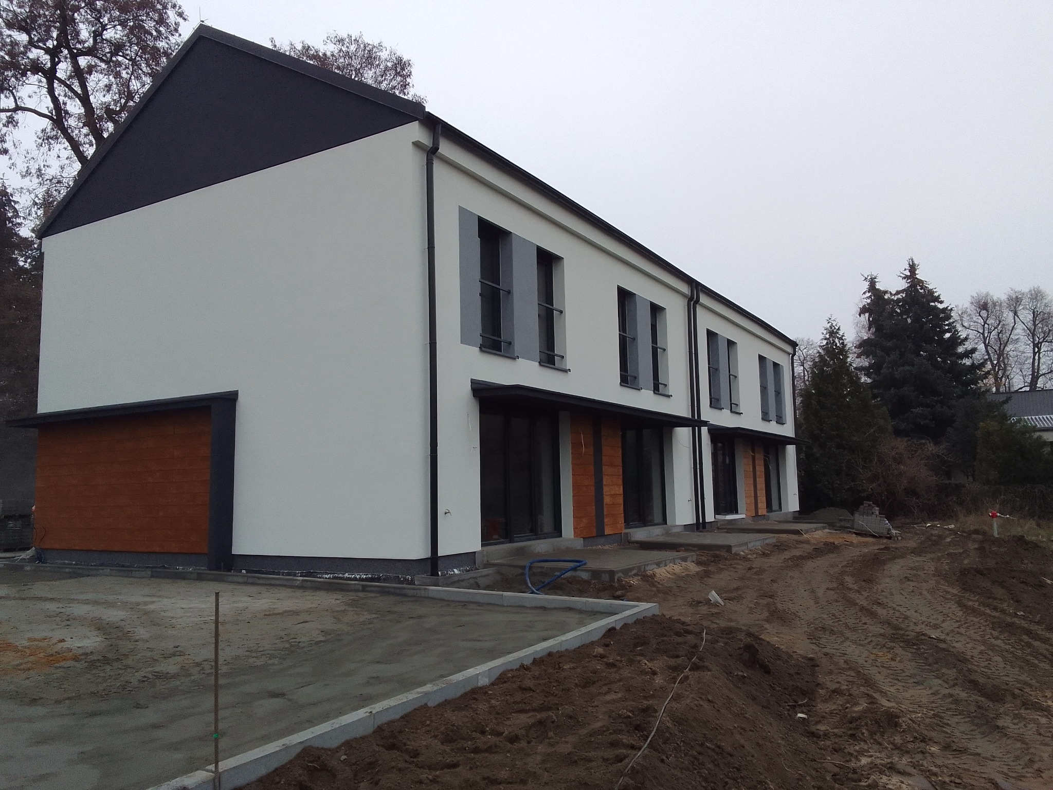 Nowoczesny szeregowy dom z białą elewacją, czarnym dachem i drewnianymi akcentami, widoczny w surowym krajobrazie przed domem, z nieukończonym chodnikiem i ziemią.
