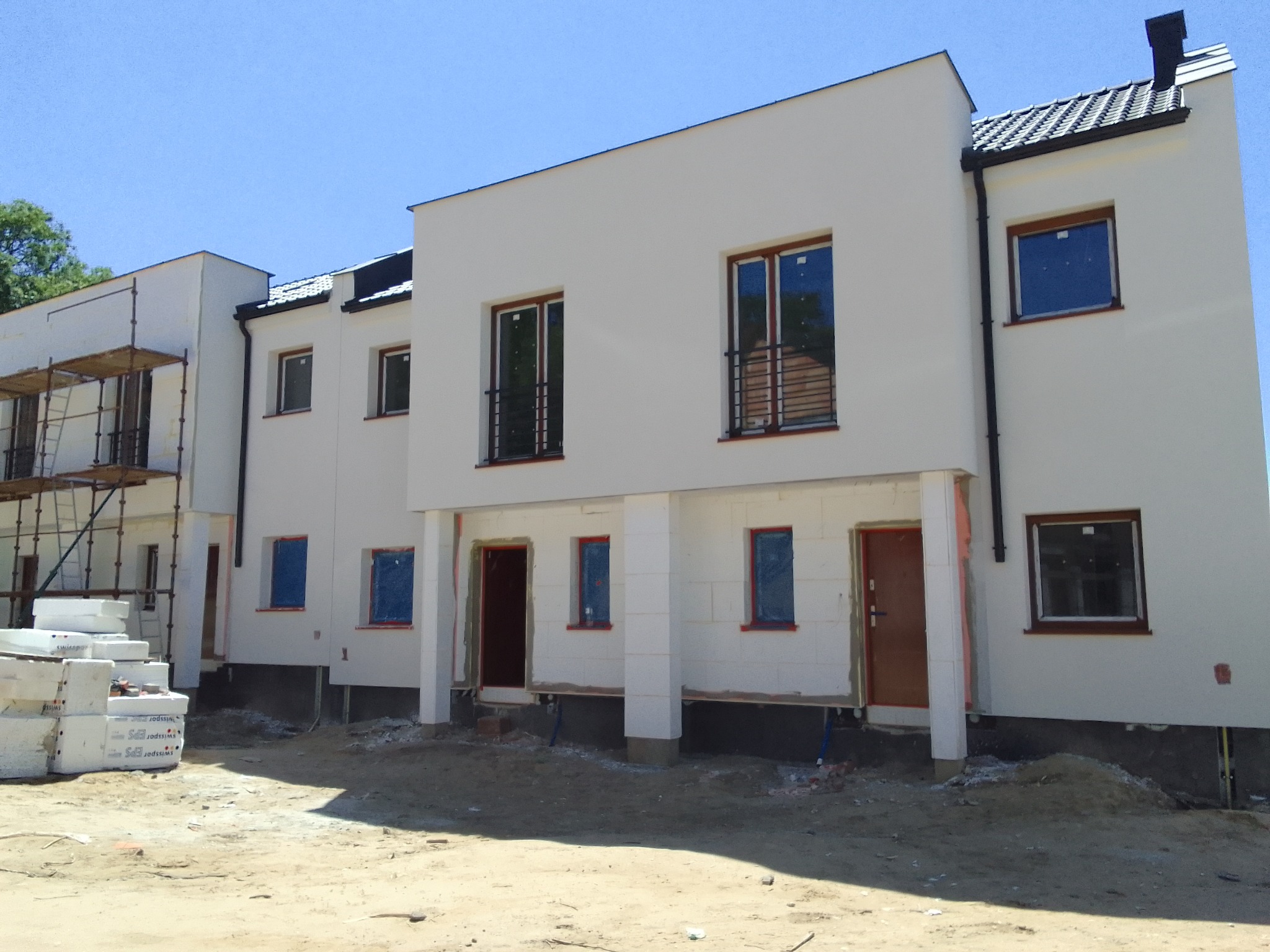 Nowe budynki w trakcie prac wykończeniowych elewacji, widoczne rusztowania, okna zabezpieczone folią, teren wokół budynków niezagospodarowany.