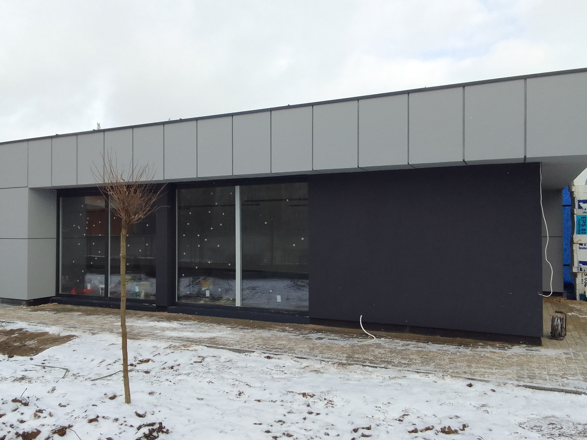 Nowoczesny budynek z szarą elewacją panelową i czarną ścianą, duże okna, zimowy krajobraz z młodym drzewkiem na pierwszym planie, palety materiałów budowlanych w tle.