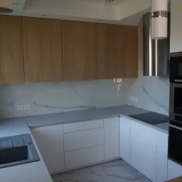 Nowoczesna kuchnia z drewnianymi szafkami górnymi, białymi szafkami dolnymi, szarym blatem i marmurowymi płytkami na ścianie, z widocznym zlewem i sprzętem AGD w zabudowie.