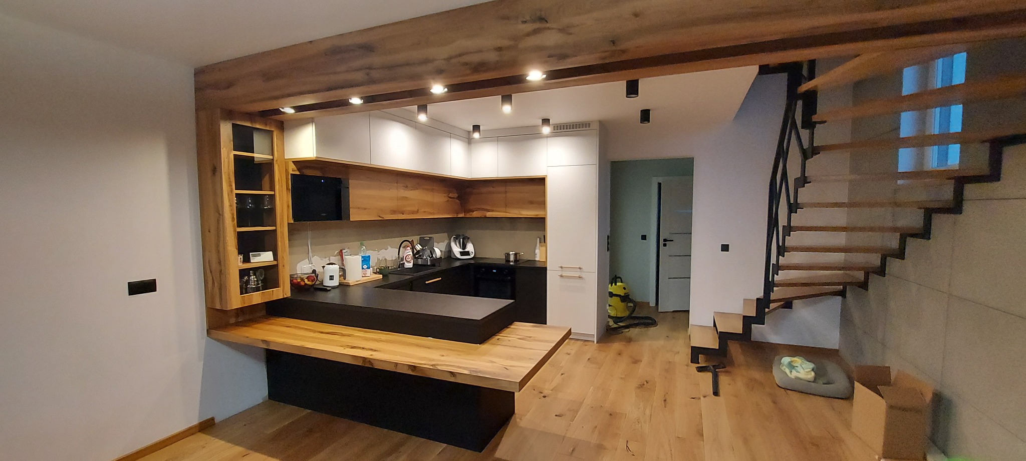 Nowoczesna kuchnia z drewnianymi akcentami: blaty z naturalnego drewna, czarne fronty szafek i białe górne szafki, oświetlenie punktowe, drewniana belka nad kuchnią, schody z drewnianymi stopniami...