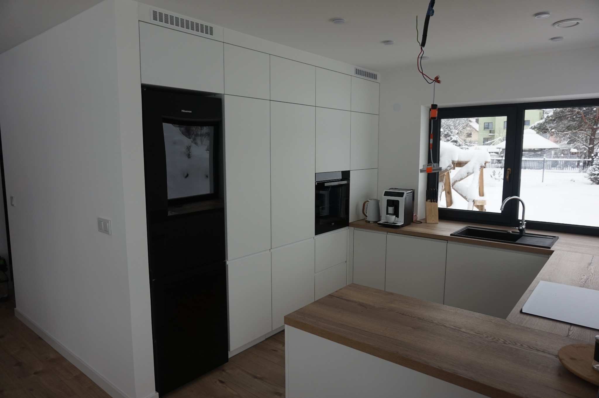 Nowoczesna, minimalistyczna kuchnia z białymi frontami szafek i drewnianym blatem, czarny sprzęt AGD, widok na zaśnieżony ogród przez okno, niedokończone oświetlenie sufitowe.