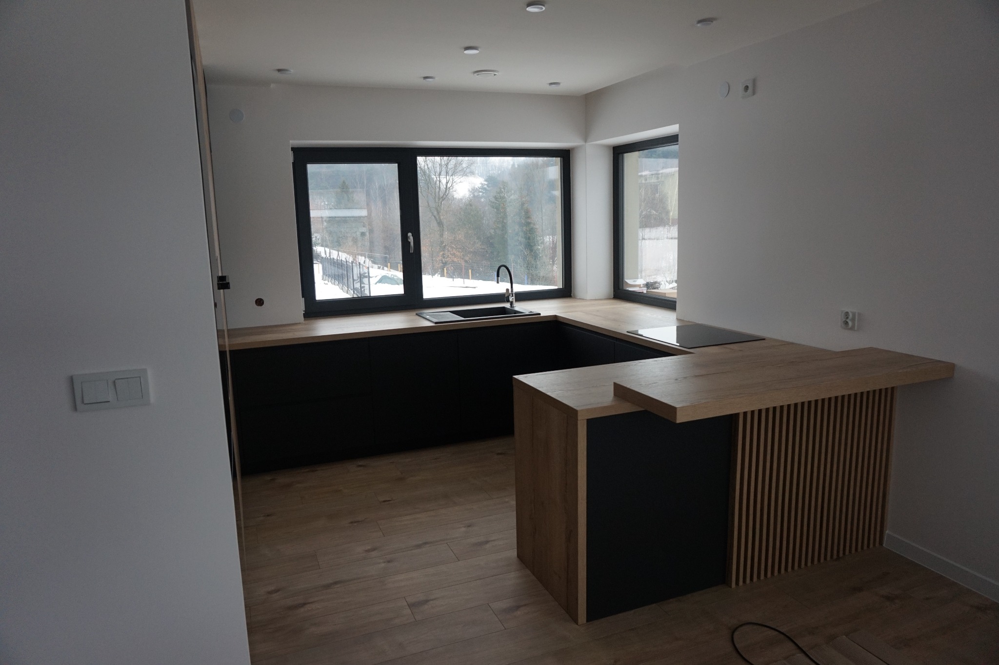 Nowoczesna kuchnia z czarnymi szafkami, drewnianym blatem i wyspą z lamelami, widok na zimowy krajobraz za oknem.