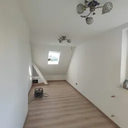 Poddasze po remoncie: białe ściany, drewnopodobna podłoga, okno dachowe i ozdobne lampy sufitowe. Na podłodze stoi odkurzacz przemysłowy.